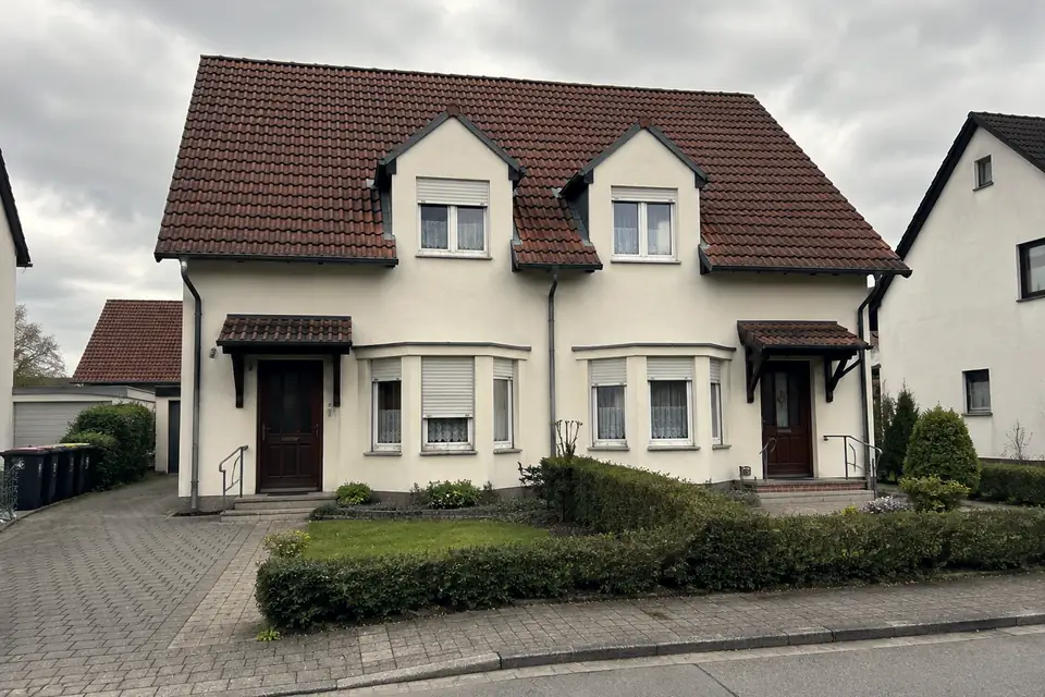Älteres Doppelhaus mit zwei Gauben, hellem Putz und rotem Ziegeldach, davor eine geschnittene Hecke und eine gepflasterte Zufahrt