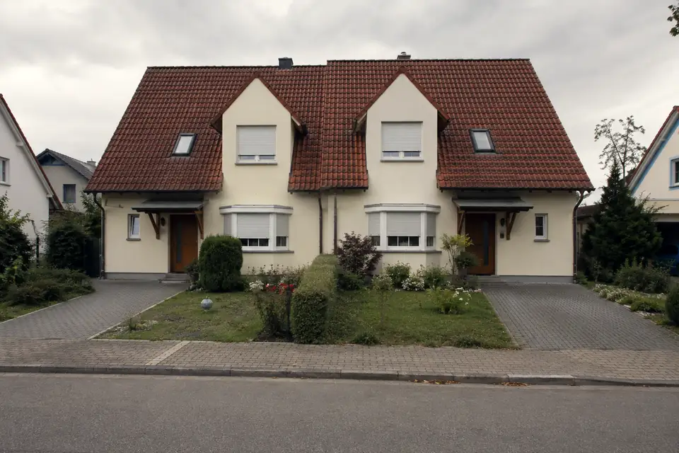 Doppelhaus jüngeren Baujahrs mit zwei Zwerchgiebeln, hellem Putz und rotem Ziegeldach, davor gepflasterte Auffahrten und ein bepflanzter Vorgarten