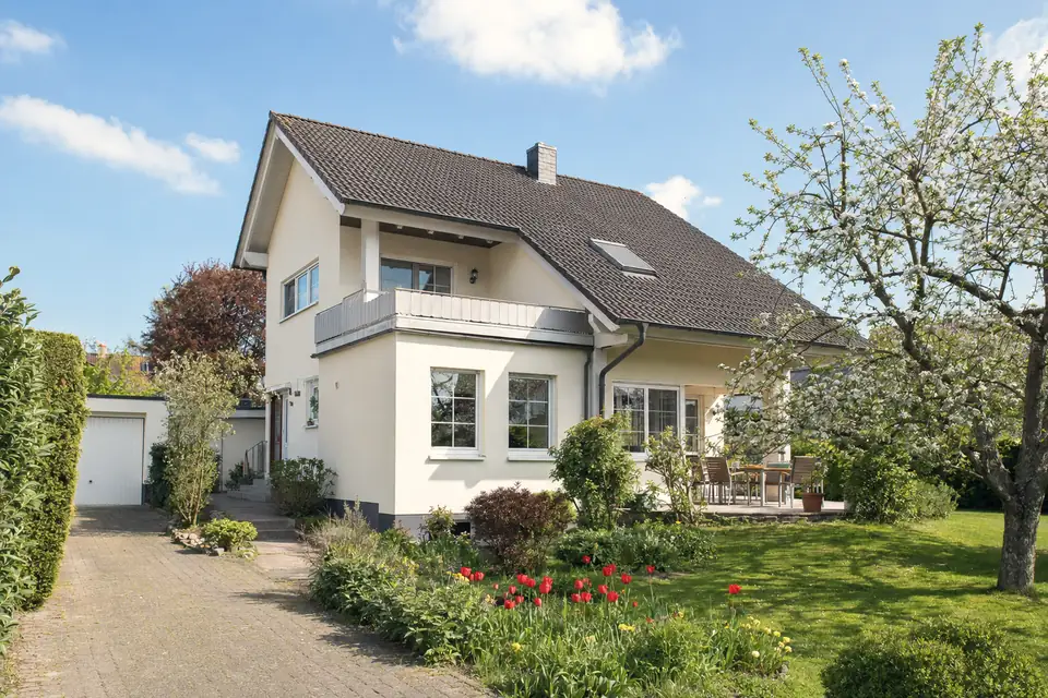 Freistehendes Einfamilienhaus mit Satteldach, Balkon und Garage, davor ein Garten mit blühendem Apfelbaum und einem Tulpenbeet