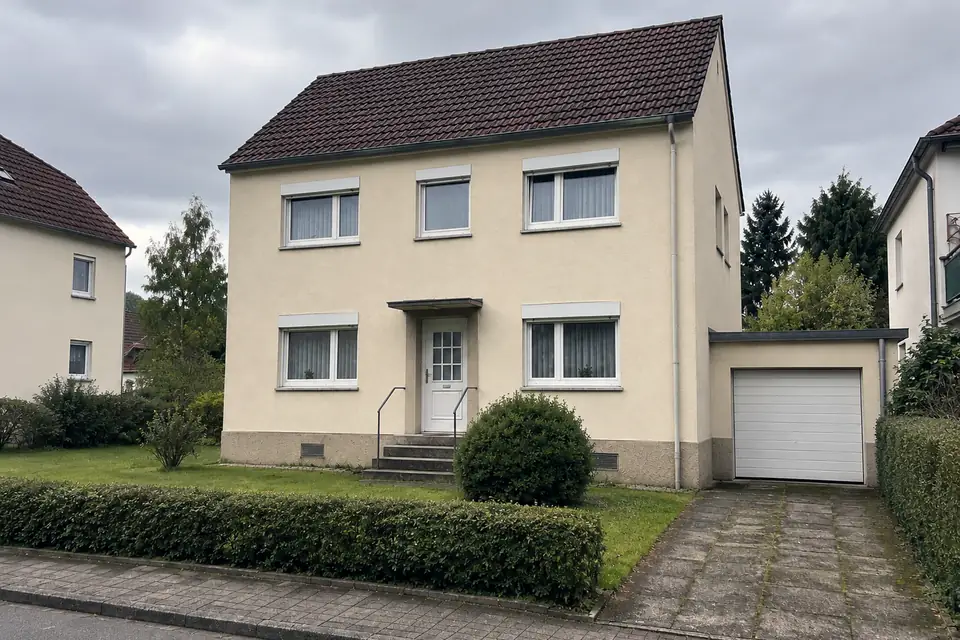 Freistehendes Einfamilienhaus mit Satteldach und angebauter Garage, hellem Putz und einer Hecke zum Gehweg